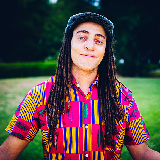 Photo of Vuli he wears a blue wool textured type flat cap facing forward, he has long brown dreadlocks that run past his chest  towards his ribs, he wears a colourful African pink, blue and yellow short sleeved shirt, he has a wry smile on his face and the background is blurry but looks like green grass and trees